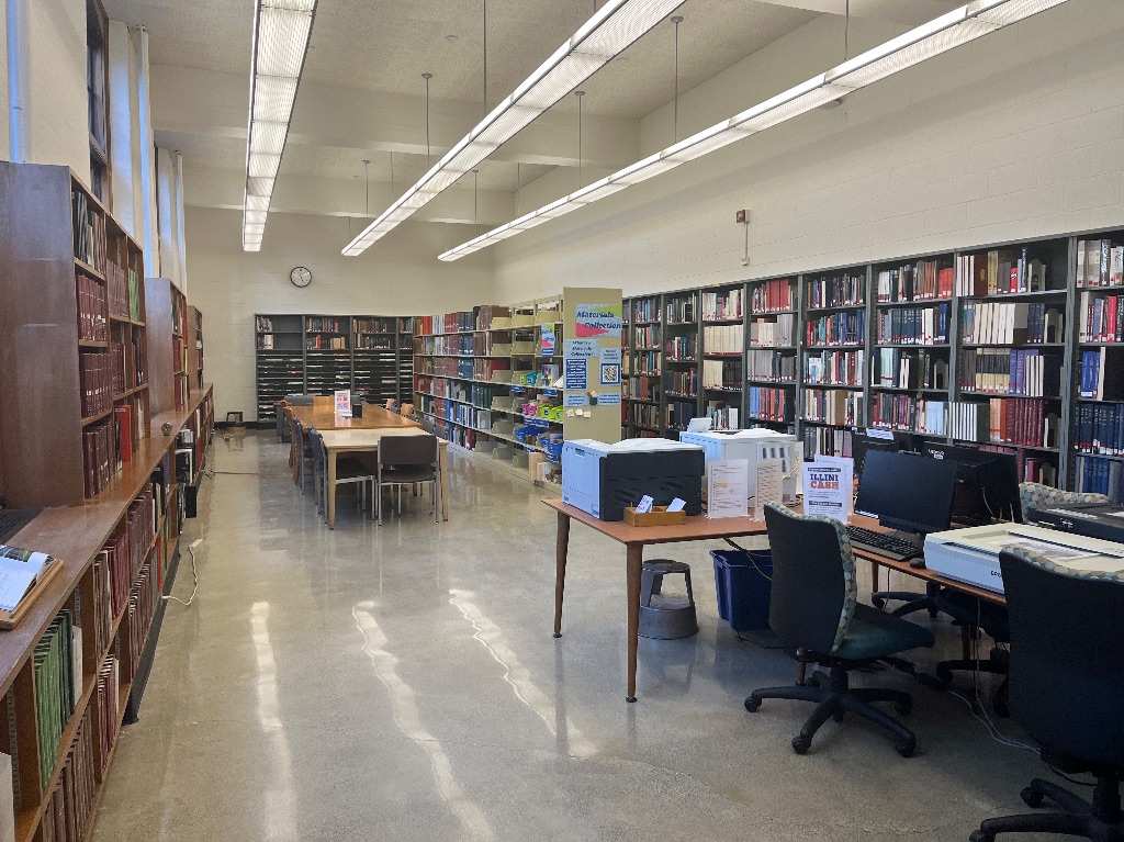 A large library room with bookshelves on every wall. there is a long table towards the back of the room and on the right side is a desk with computers and printers.