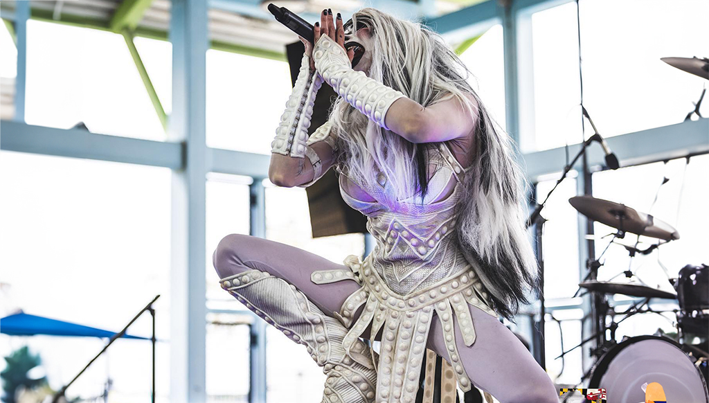 A performer is captured mid-song on stage, microphone in hand. They are attired in a striking white outfit, adorned with spikes, and a long white wig. The outfit is illuminated by a purple light. The performer is in a dynamic pose, kneeling on one knee with the other leg extended. The backdrop features a blue metal structure and a drum set.