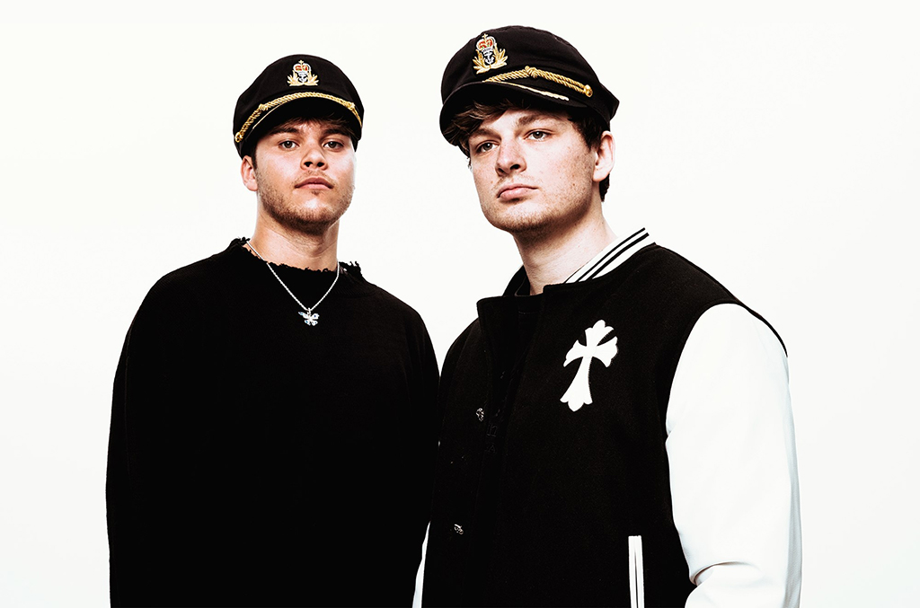 Two individuals stand side by side, both donning black captain's hats adorned with gold badges. The person on the left is attired in a black shirt, complemented by a silver necklace. The individual on the right sports a white shirt, distinguished by a black cross emblazoned on it. They are set against a plain white backdrop.