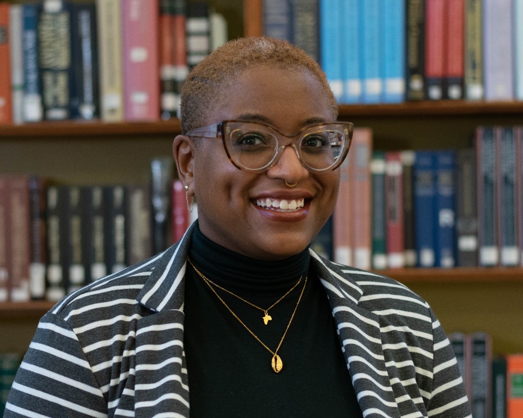 A black woman in large tortoise glasses and close cropped hair stands in front of a library book shelf. She is wearing a black turtleneck and a black and white striped blazer with two small gold necklaces. 