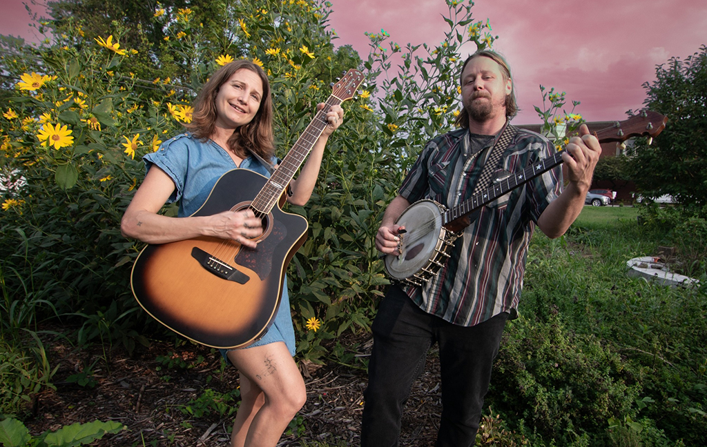 Two individuals are standing in a garden, each holding a musical instrument. The person on the left is attired in a blue dress and holds a guitar. The individual on the right is wearing a plaid shirt and holds a banjo. They are surrounded by a lush garden filled with yellow flowers and greenery under a cloudy sky with diffused lighting.