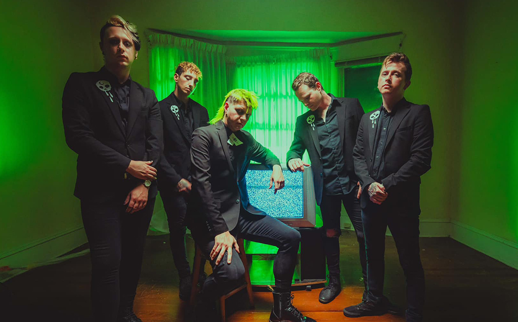 In a room bathed in the soft hues of green, six individuals stand together, their black suits a stark contrast against the backdrop. Each suit is adorned with a white flower, adding a touch of elegance. One individual breaks the standing formation, comfortably seated on a blue chair patterned in white. The ensemble exudes an air of sophistication and unity.