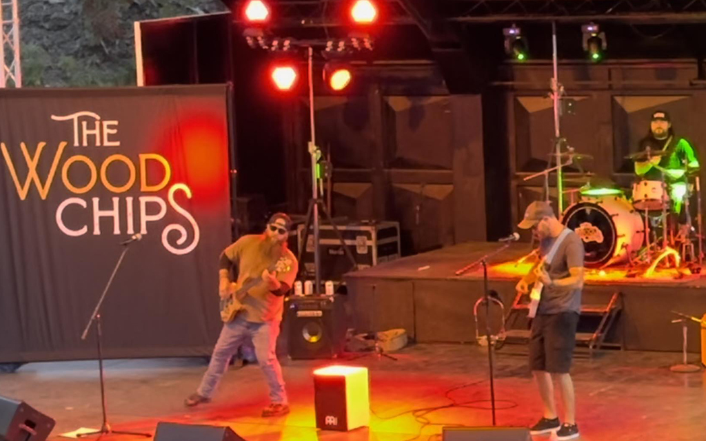 The scene captures a band performance on stage. The backdrop is black with “The Wood Chips” written in white. Three band members are present, each engaged with their instruments: drums, guitar, and bass. They are all casually dressed and wearing baseball caps.