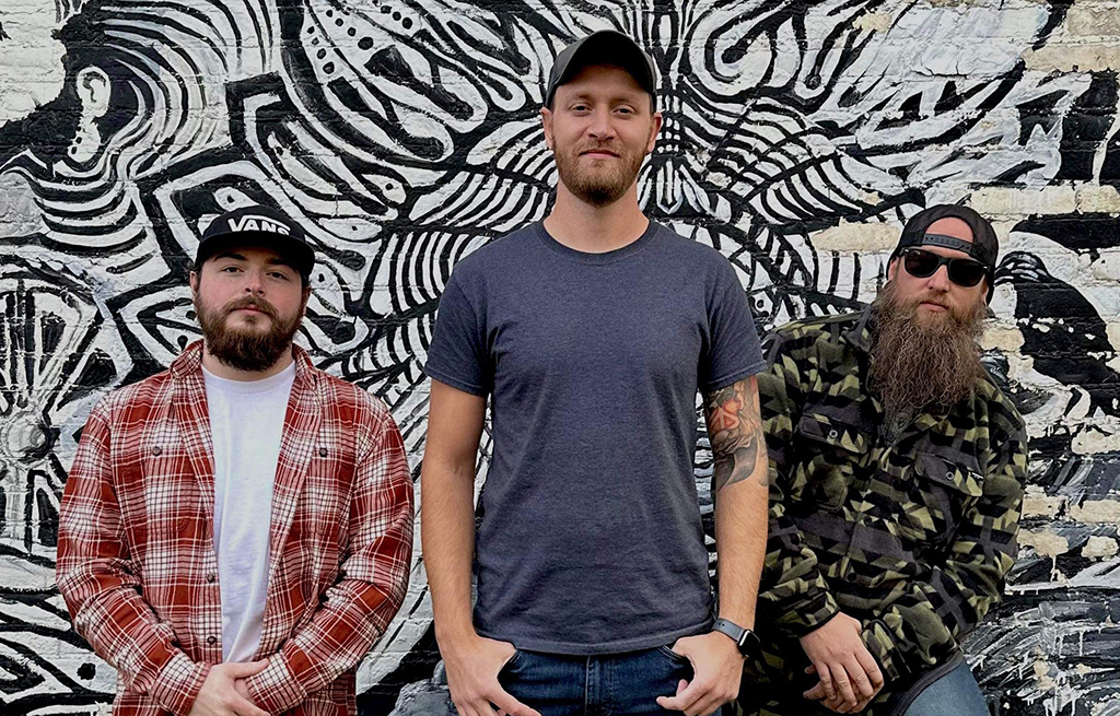 Three individuals are standing in front of a mural that is black and white with abstract designs of swirls and lines. The person on the left is clad in a red and white plaid shirt. The individual in the middle is wearing a gray t-shirt. The person on the right is donned in a green camouflage jacket.