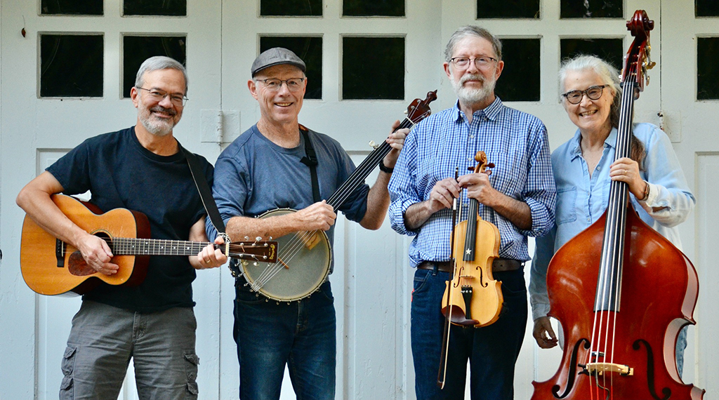 Four musicians stand smiling and looking at the camera holding their string instruments. The background appears to be the outside of a building with large white doors with windows on the top.