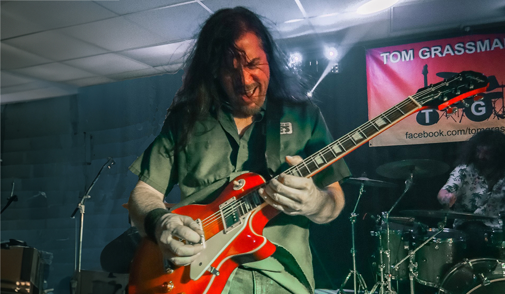 A person is captured in the midst of a performance on stage, their passion evident in the way they hold a red electric guitar. Their attire is a casual green shirt, which hangs loosely over their frame. Their long, dark hair cascades down their back, adding to the overall rockstar aesthetic. In the background, a drum set waits silently for its turn, and a banner bearing the name “Tom Grassman” hangs proudly.