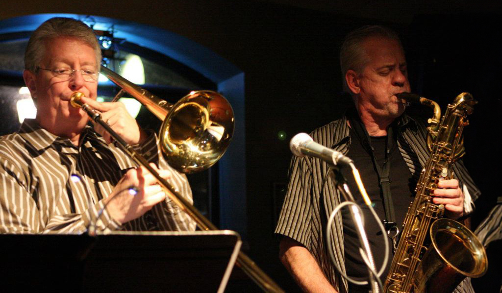 Two musicians are captured in the midst of a performance on a stage. The individual on the left is engaged with a trombone, while the one on the right is playing a saxophone. They are both dressed in black and white striped shirts. The stage setting is dark, illuminated by stage lighting.