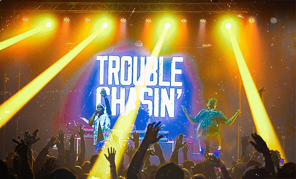 The image captures a vibrant concert scene. Two performers command the stage, bathed in the glow of yellow and orange lights. One is clad in a blue jacket, while the other sports a green shirt. The large screen in the background displays the words “Trouble Chasin’” in bold white letters. In the foreground, the audience is a sea of raised hands, their enthusiasm palpable even in stillness.