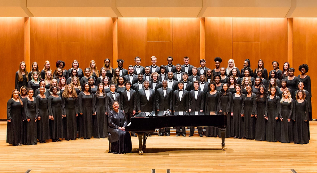 A group of individuals are on a stage, each donned in black robes with white collars. Some hold black folders. A piano is positioned in front of them, and a conductor stands to the side.