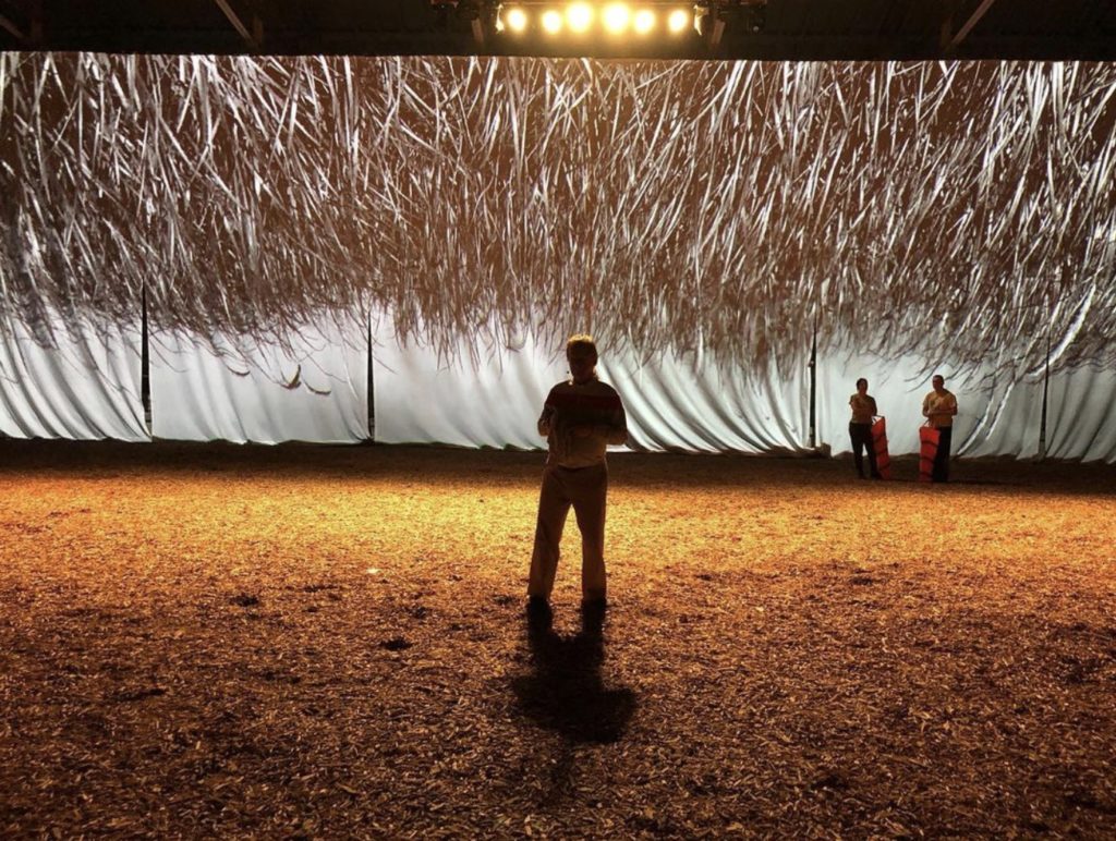 a man stands in the center of the stock pavilion. The screen is lit up behind him showing what appears to be grass from the top. 