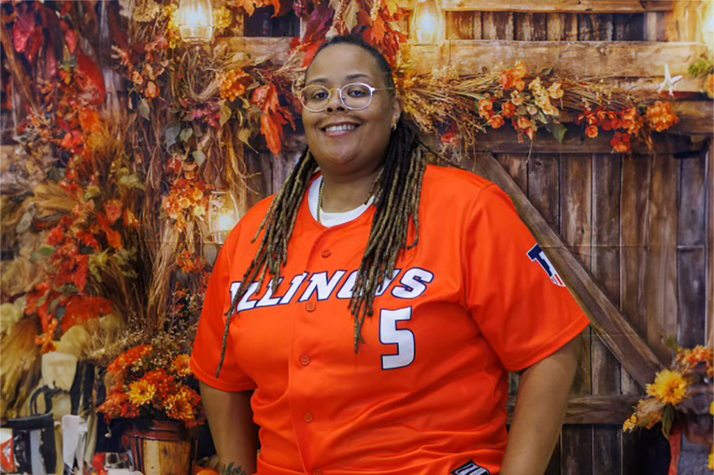 A person stands in front of a wooden wall adorned with autumn leaves and flowers. They are dressed in an orange baseball jersey with the word “Illinois” and the number “5” on it. The background features a wooden wall decorated with autumn leaves and flowers, and a wooden bench is visible on the right side.