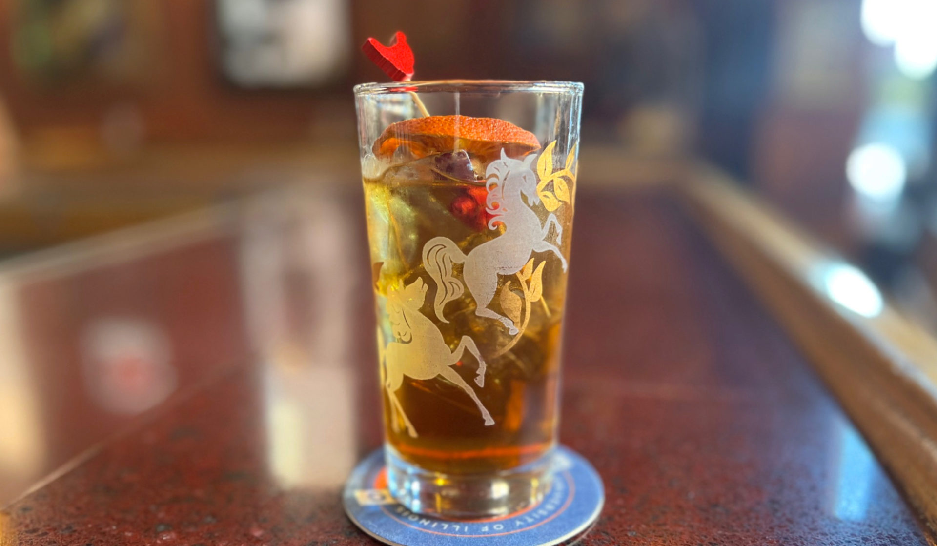 Watson's cocktail, a twist on a Vieux Carre drink in a glass etched with flying ponies, garnished with an orange and two rosebuds on a rooster toothpick.
