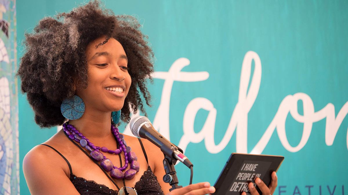 A Black woman in a black tank top and chunky purple necklace is standing at a microphone, reading from a card.