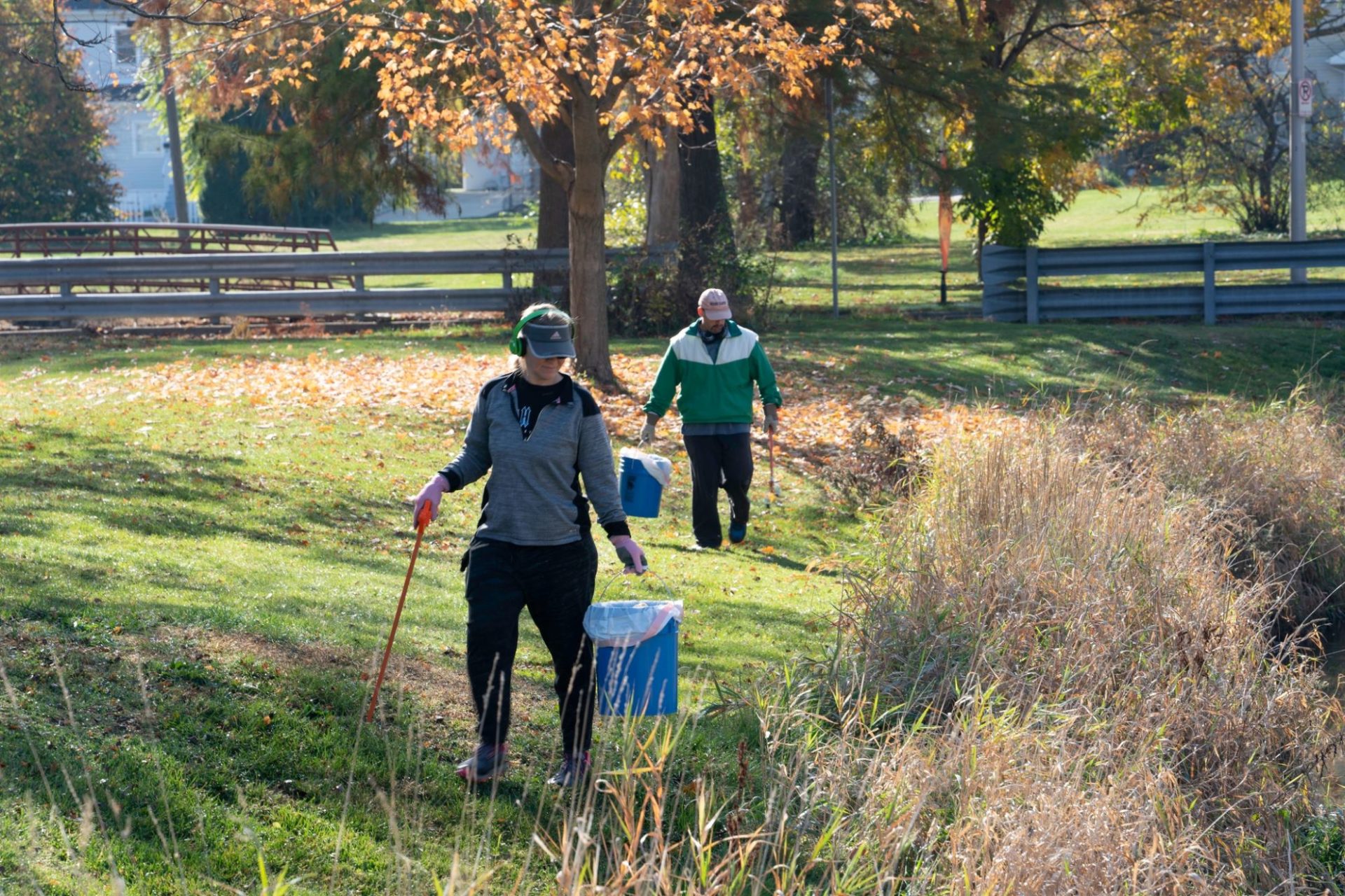 Two people walk along a creek bed carrying buckets and sticks for picking up litter.