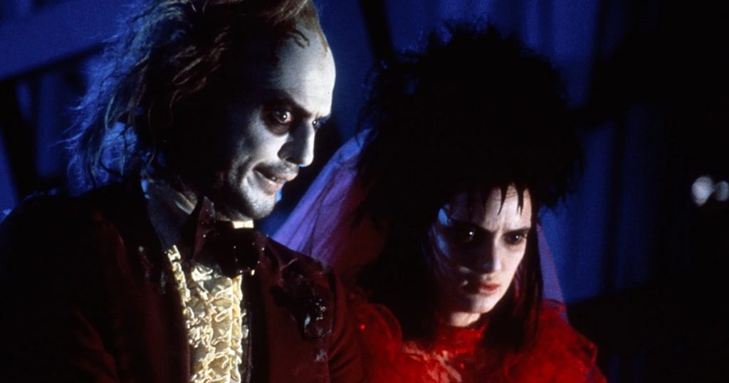 Screenshot from Beetlejuice, with the title character standing alongside Winona Ryder, with spiked black hair and a red dress.