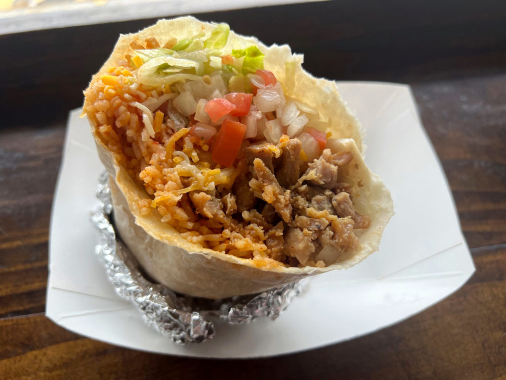 A burrito from Cactus Grill with honey chicken, orange rice, onions, tomato, lettuce, and cheese in a flour tortilla wrapped with tin foil in a white paper basket.
