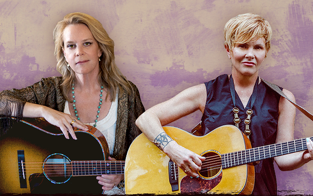 Two individuals are captured in the image, both engrossed in playing their guitars. The individual on the left is attired in a black top adorned with a floral pattern and a turquoise necklace. The individual on the right is seen in a black vest with gold buttons, their arm showcasing a tattoo. The backdrop of the image is a solid purple.