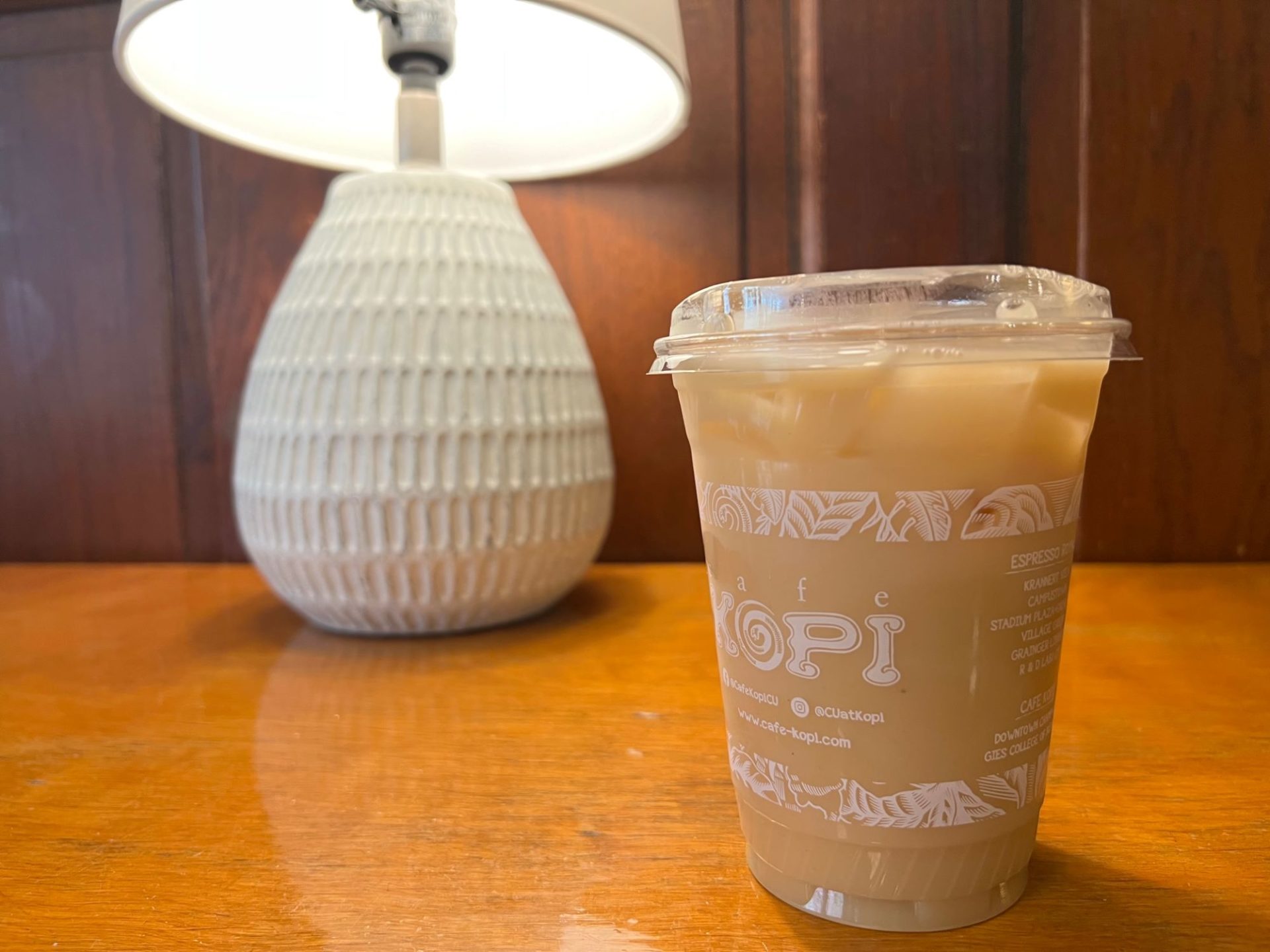 A plastic cup is filled with a light brown liquid, sitting on a light wood table with a small white lamp.