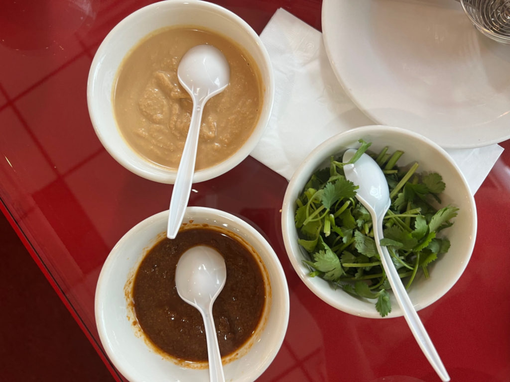 Three white porcelain bowls with white plastic disposable spoons. The top left has peanut sauce, the bottom left has chacha sauce, and the right has fresh chopped cilantro.