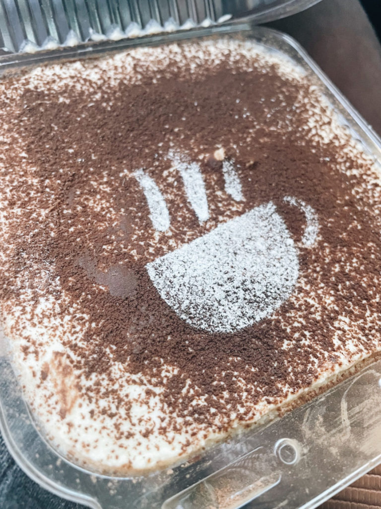Filling a clear clamshell container is tiramisu. It fills the container, showing a top layer of dusted cocoa powder. A powdered sugar coffee cup with three “steam” lines is dusted atop that.