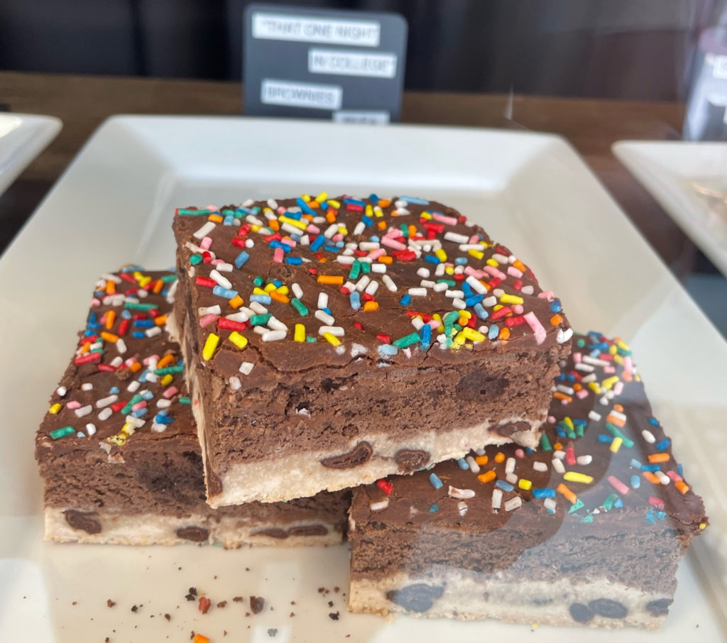 On a white square plate is a pile of three brownies. The individual layers show a chocolate chip cookie base, topped with a brownie layer and topped with rainbow sprinkles.