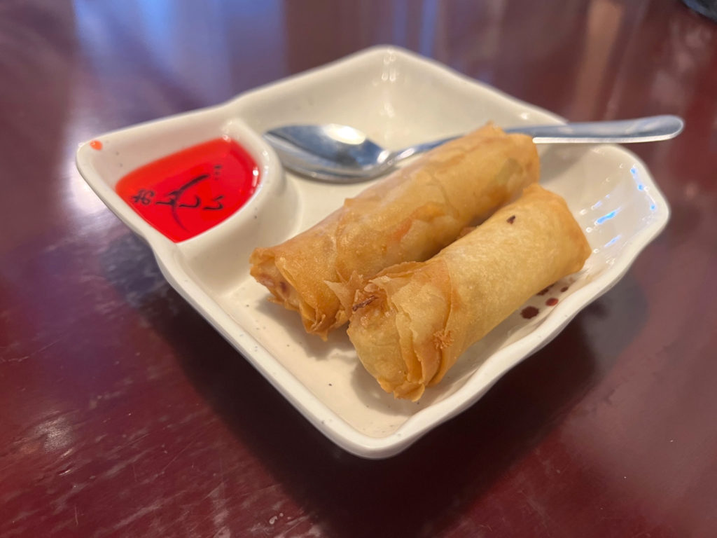 Two egg rolls on a small plate with bright red dip sauce in its own compartment and a silver spoon.