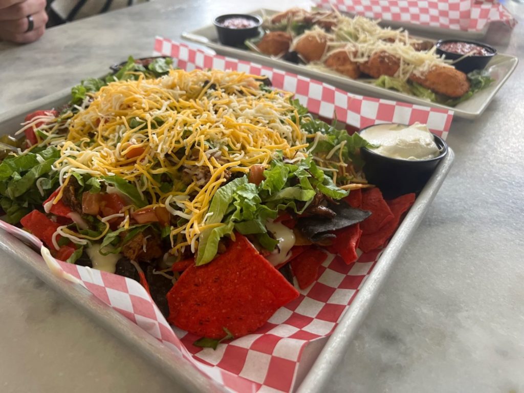 A tray of nachos with multi-colored chips, cheese sauce, lettuce, tomato, and shredded cheese.