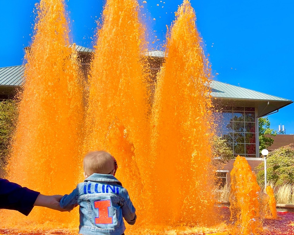a baby wearing a jean jacket with an orange. She sands in front of a water fountain with bright orange water and a bright blue sky.