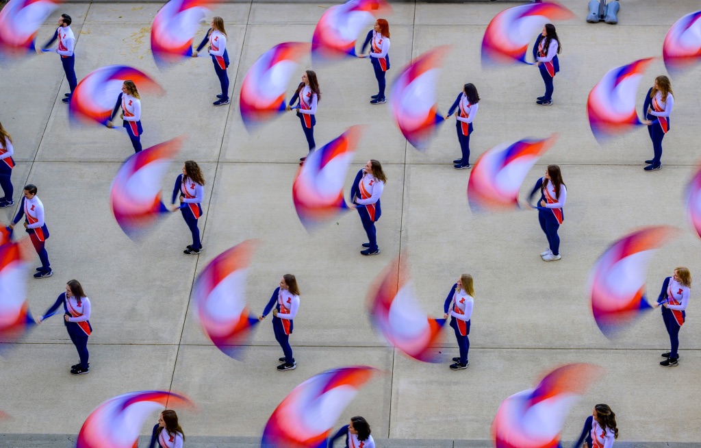 An arial shot of flag twirlers standing in straight lines wearing the same blue pants and white and orange jackets and twirling their flags which are blurry