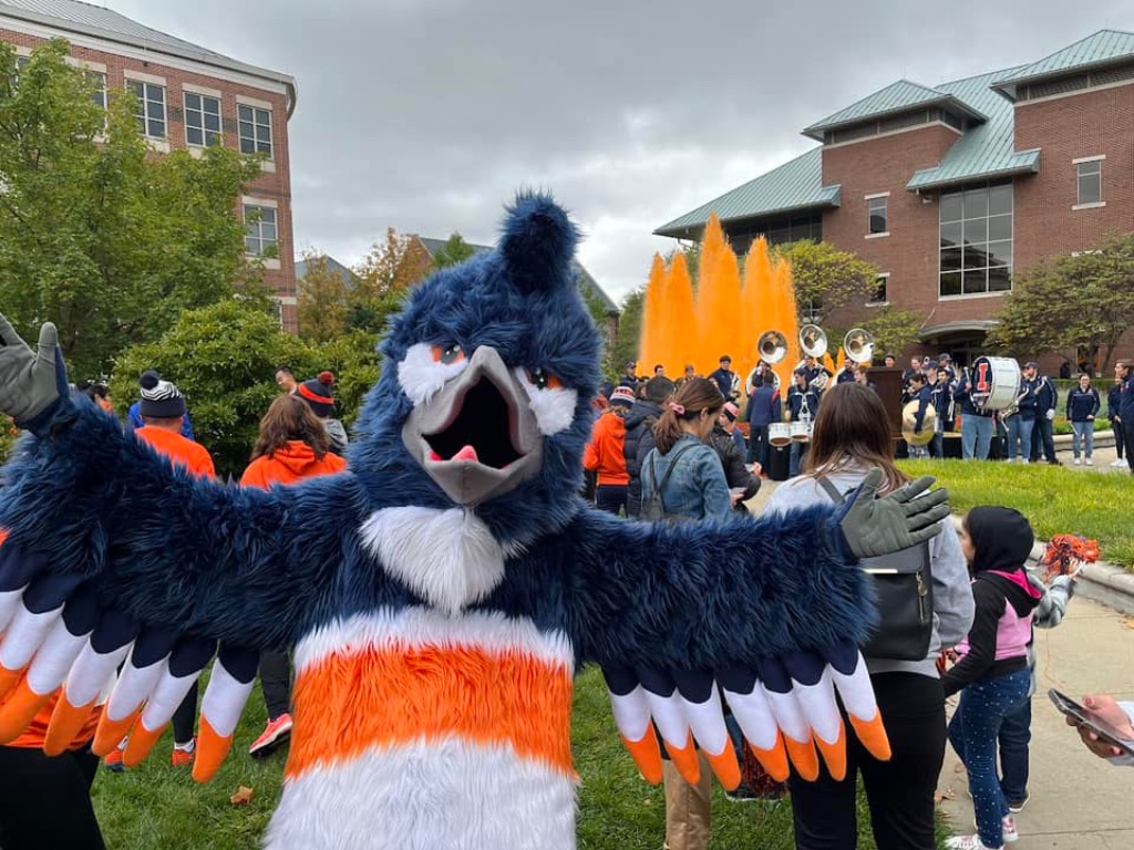 A Kingfisher mascot is spreading its wings in front of a fountain with orange water and a group of people behind it. The mascot is a bird with a large grey beak blue, white, and orange feathers, and a blue and white body with an orange belt.