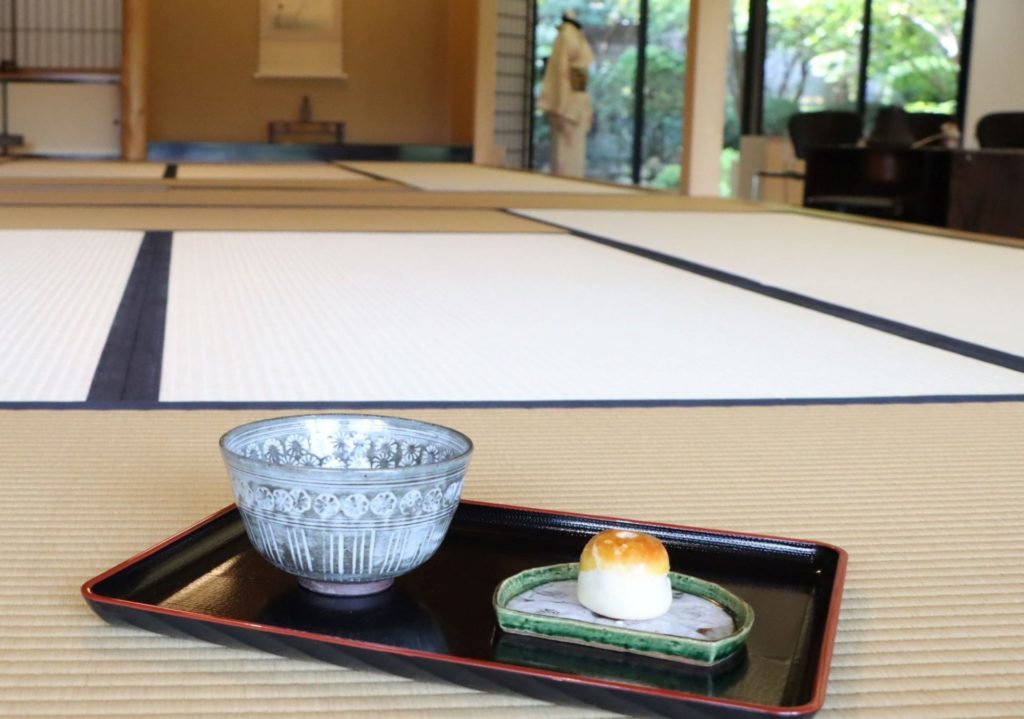 A bowl of matcha and a wagashi sweet are placed next to each other on a black tray. The tray is resting on a tatami mat at Japan House.