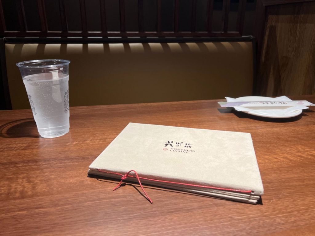 A table with a menu, a plastic cup of water, a white plate with a white paper napkin, and paper enclosed chopsticks.
