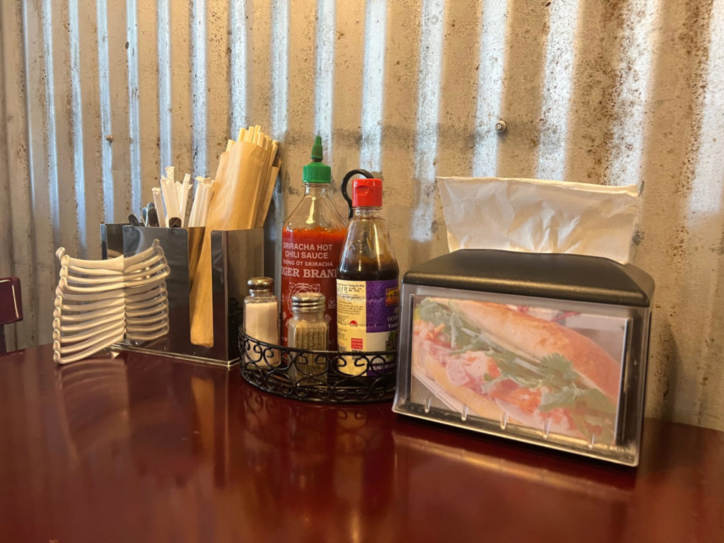 On the table, there is a utensils holder with white stacked soup spoons, wrapped straws, chopsticks in brown paper, a little black carrier of sriracha, soy, sesame oil, salt, and pepper shakers beside a napkin dispenser with a banh mi photo on it.