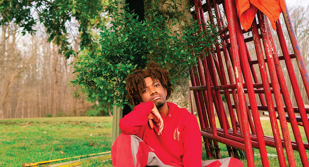 An individual is seated under red steel grates in a park setting. They are dressed in a red sweatshirt and sweat pants The grates, made of red painted steel are leaning on a tree with green leaves. The surrounding area is a grassy field dotted with trees.