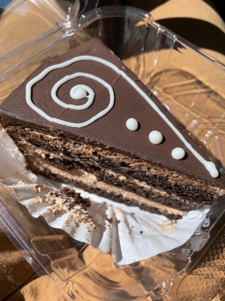 Rick’s chocolate mousse cake: A triangular slice of chocolate cake sits in a wrapper in a clear clamshell container. The cake has a white swirl and three small dots piped on top of a smooth layer of chocolate.