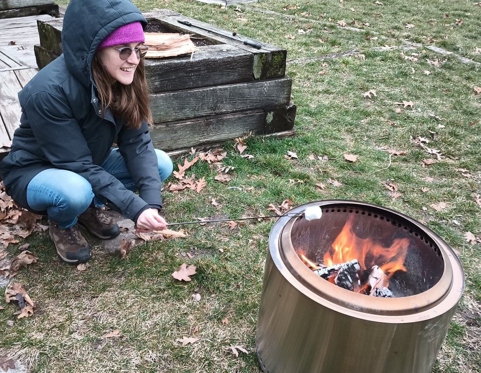 A girl with long hair, purple stocking cap, and gray coat is kneeling near a firepit, roasting a marshmellow.