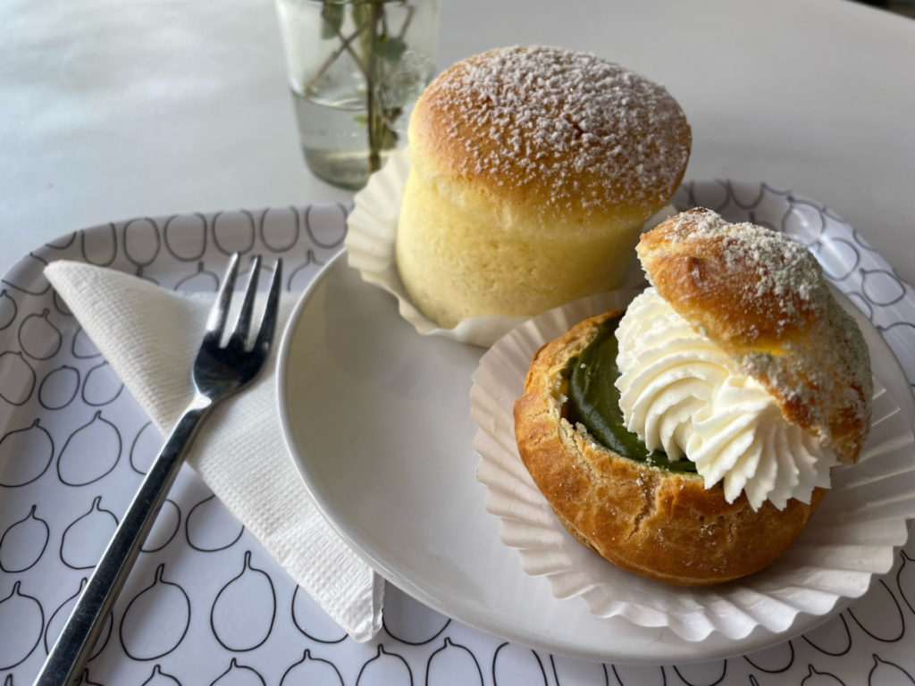 On a plate sits a Japanese cheesecake, a pale yellow dome-cylinder. It is dusted with powdered sugar. Next to that is a matcha shu cream puff: a round crust topped with green matcha cream, dolloped with whipped cream, and topped with the other have of the round crust. A fork is to the left of the sweets.