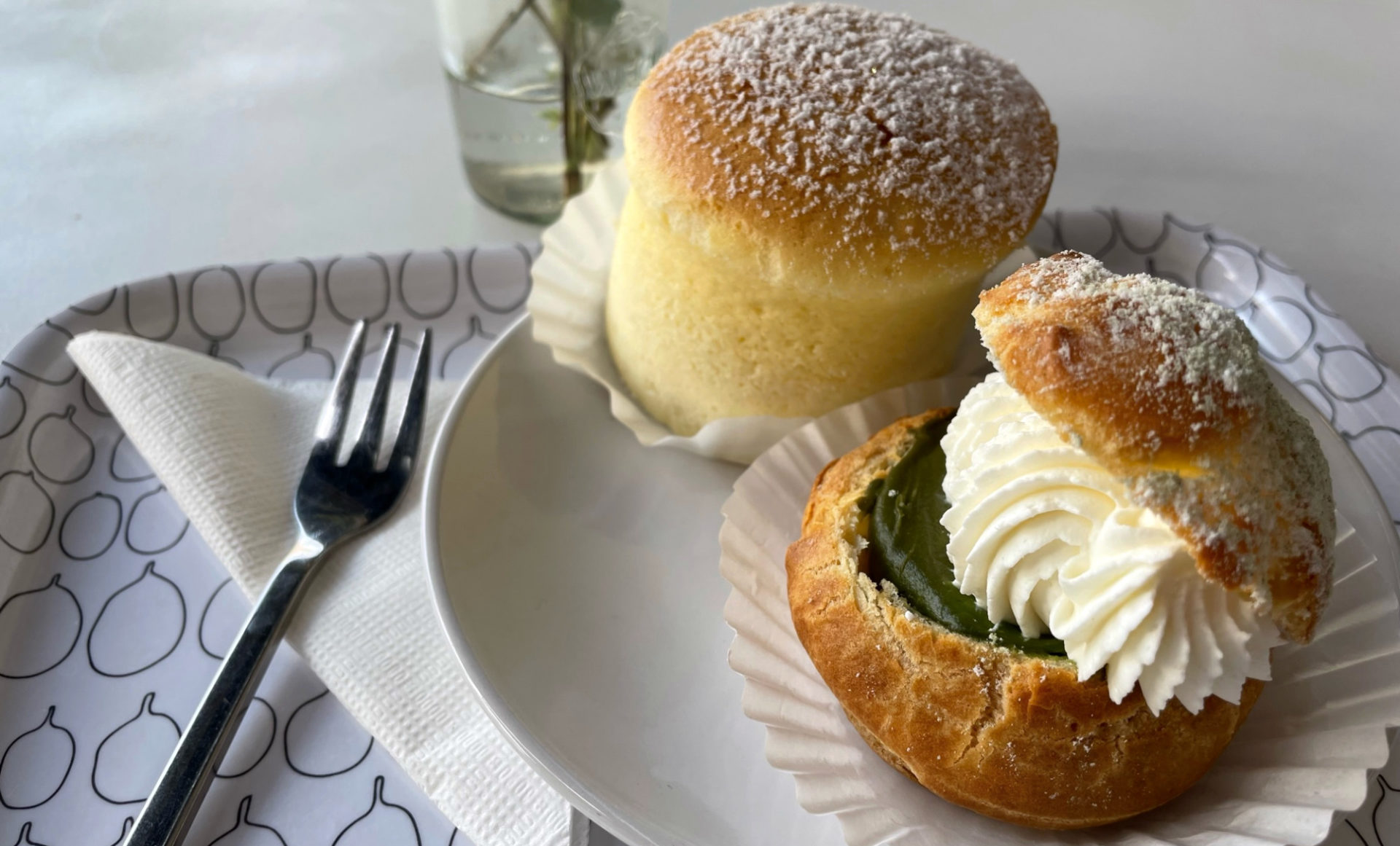 On a plate sits a Japanese cheesecake, a pale yellow dome-cylinder. It is dusted with powdered sugar. Next to that is a matcha shu cream puff: a round crust topped with green matcha cream, dolloped with whipped cream, and topped with the other have of the round crust. A fork is to the left of the sweets.