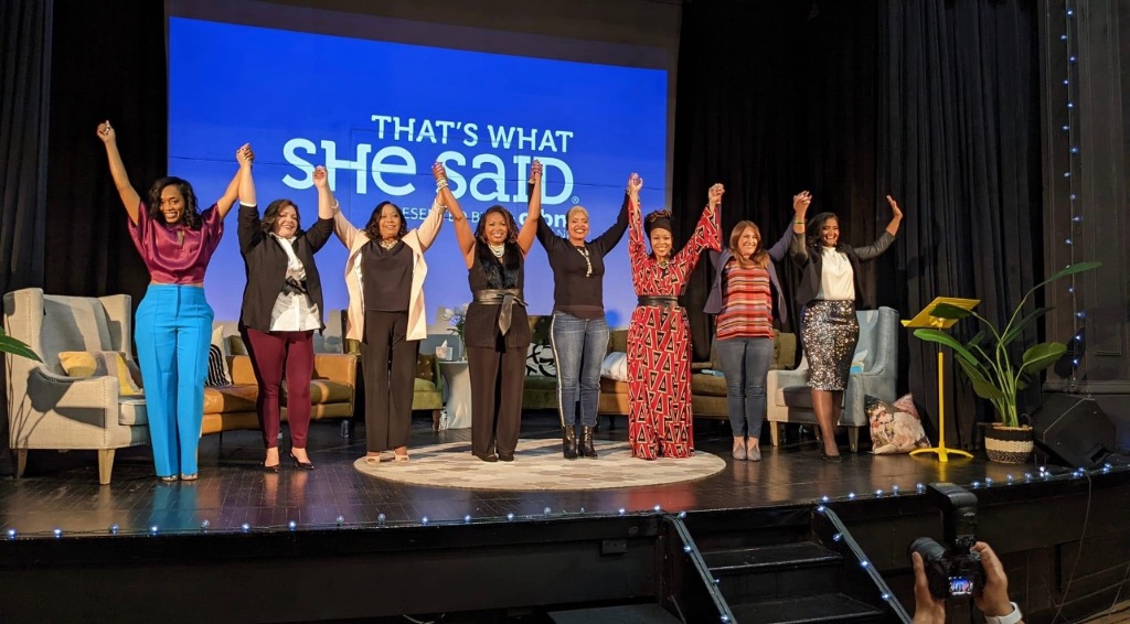 Eight woman stand on a stage with their hands together and their arms raised, about to bow. They are in front of a blue screen that says That's what she said.