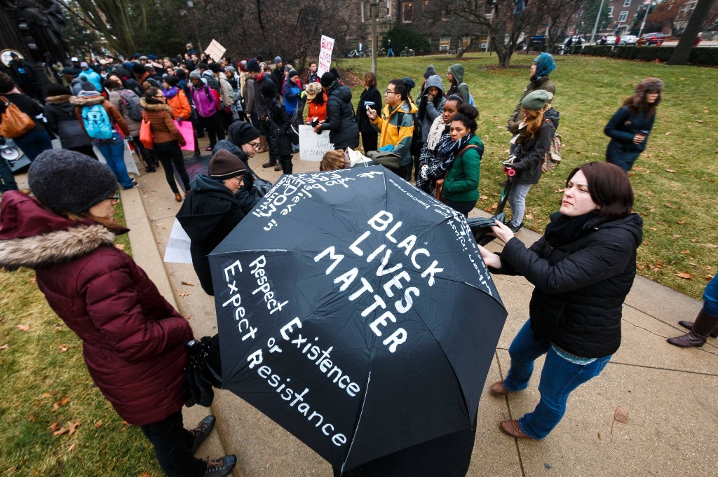 A group of people in coats stand on a sidewalk on a grey day. In the middle of the picture is a black umbrella which has "Black Lives Matter Respect Existence or Expect Resistance" written in white letters on it. 