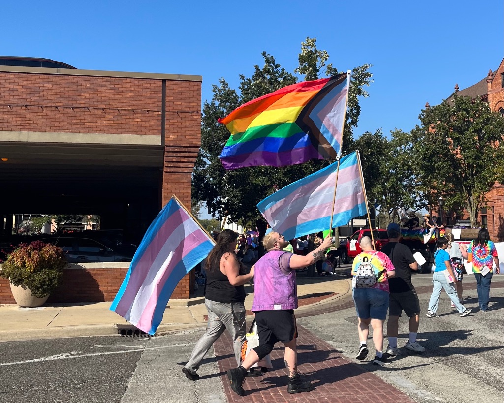 Three people walking in the street holding a variety of pride flags there is a blue sky in the background.