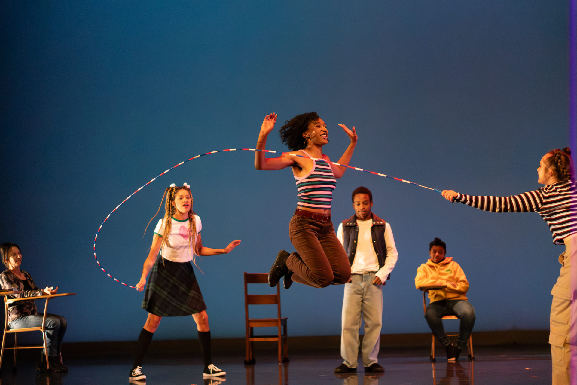 Dancers on stage against a blue backdrop. A black dancer is jumping rope as two other dancers swing the rope and others sit at desks surrounding the stage