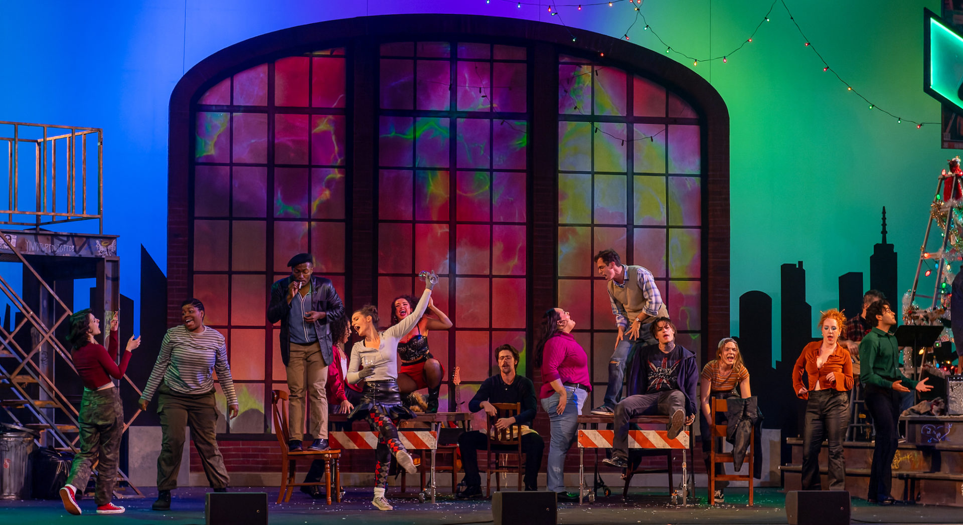 A colorful large arched window serves as the background while the actors from rent appear on stage singing