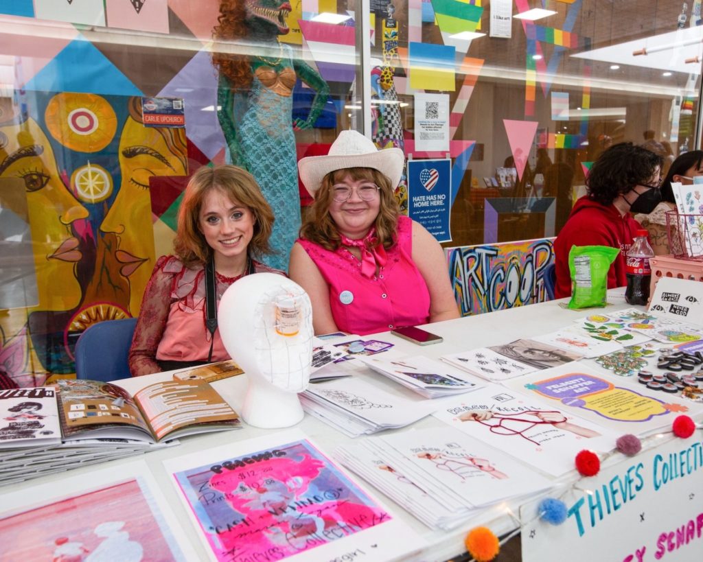Two women sit behind a table at small press fest, one wearing hot pink and a white cowboy hat