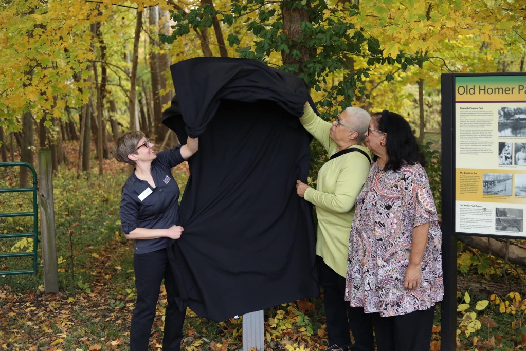 three women unveil a black cloth over a new trail sign. Two on the right are black and wearing long sleeves and pants and the woman on the right is white with short brown hair. They are standing in front of fall trees with yellow leaves. 