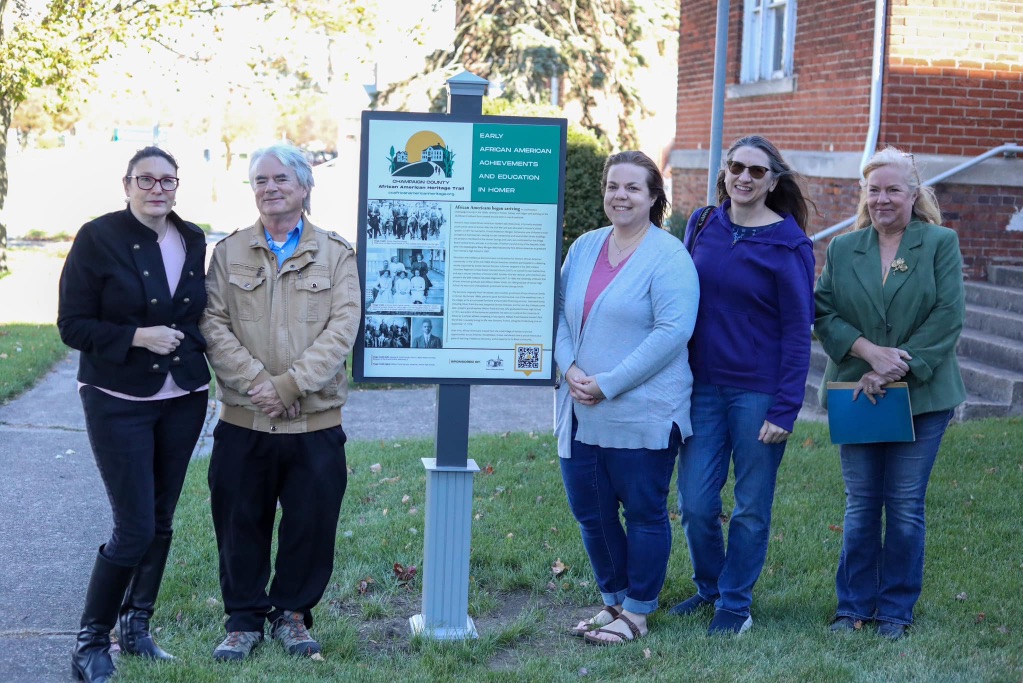 a group of white people gather around a sign from the African American Heritage Trail in Homer, IL they are wearing long sleeves and pants and standing near a brick building. 