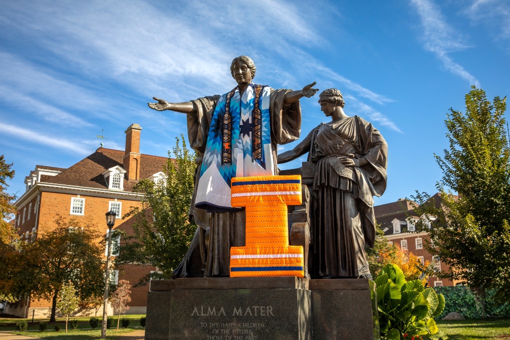 The Alma Mater statue stands with arms outstretched wearing a traditional robe in light blue, dark blues, and white colored stripes. There is also a large orange I decorated in white and dark blue stripes . There is a big blue sky above.