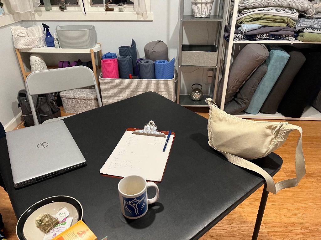 A table sits in a yoga studio. The table has a computer, clipboard, small white bag, and tea. Against the wall there are yoga mats, blocks, and blankets folded and stacked. 