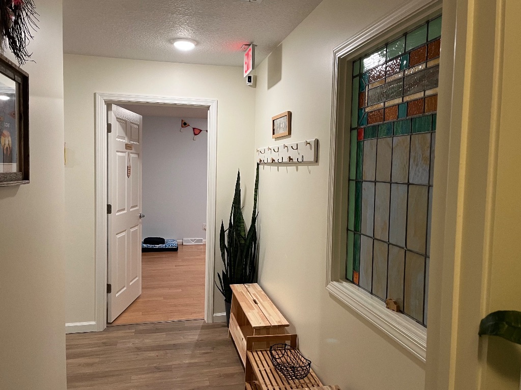 A white hallway with a door at the end. in the hallway there is a stained glass window on the right, then a bench and plant next to the doorframe. 