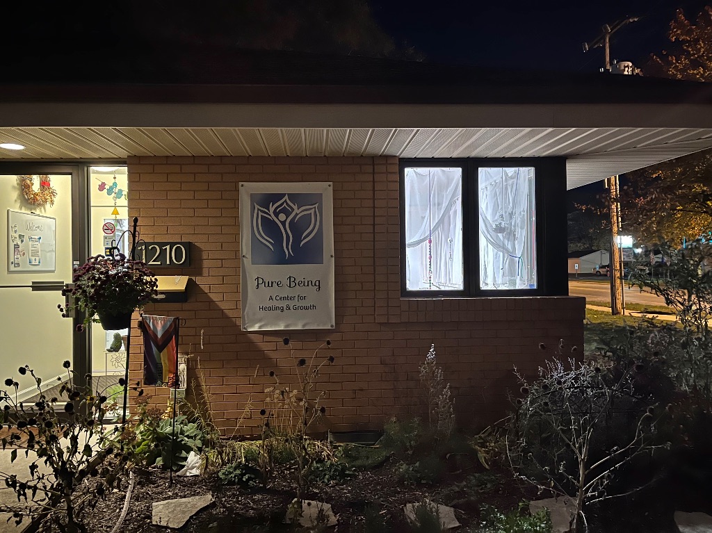 The outside of pure being. it is a one story brick building at night with the lights on inside. There are plants and bushes in front and a white sign with blue letters that says pure being.
